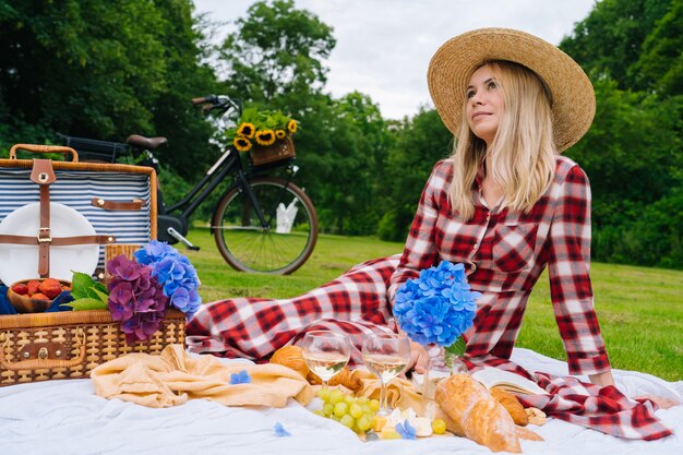 Fille en robe à carreaux rouge et chapeau assis sur une couverture de pique-nique en tricot blanc, lire un livre et boire du vin. Pique-nique d'été aux beaux jours avec pain, fruits, bouquet de fleurs d'hortensia. Mise au point sélective