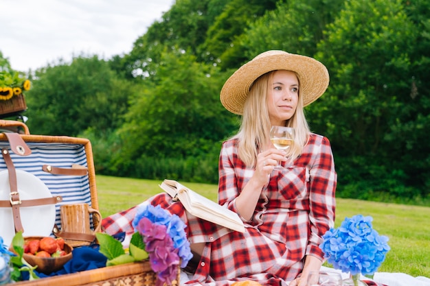 Fille en robe à carreaux rouge et chapeau assis sur une couverture de pique-nique en tricot blanc, lire un livre et boire du vin. Pique-nique d'été aux beaux jours avec pain, fruits, bouquet de fleurs d'hortensia. Mise au point sélective.