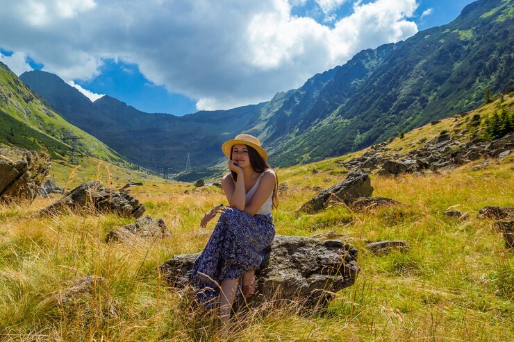Photo fille en robe et chapeau de paille est assise sur un rocher dans le contexte de hautes montagnes.