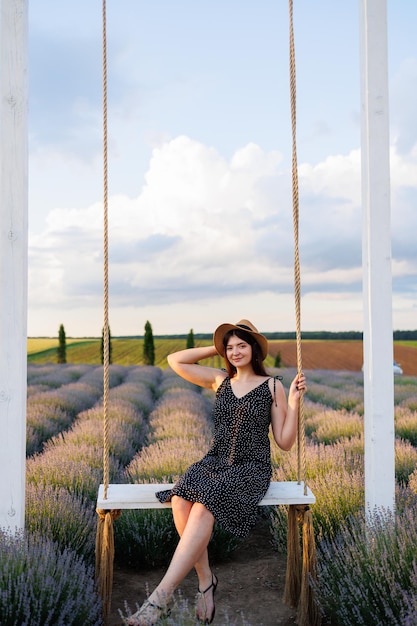 Fille en robe et chapeau posant assis sur une balançoire dans un champ de lavande