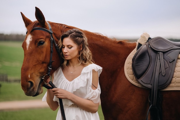 Une fille en robe d'été blanche se tient à côté d'un cheval brun dans un champ en été