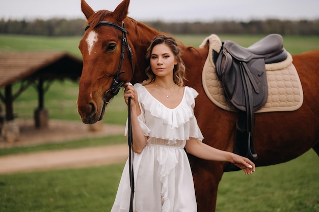 Une fille en robe d'été blanche se tient à côté d'un cheval brun dans un champ en été