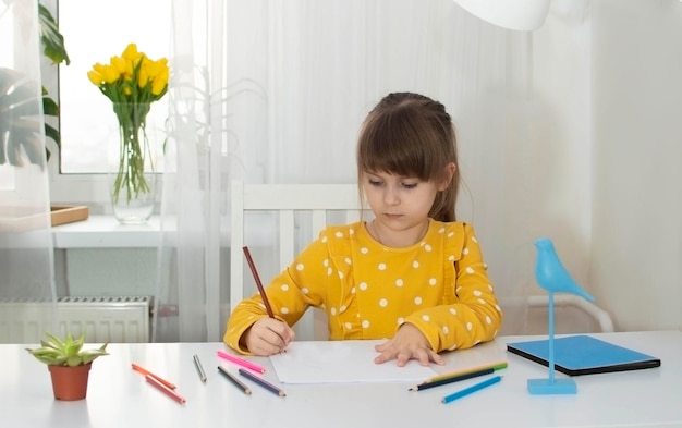 Une fille en robe jaune dessine dans sa chambre à son bureau avec des crayons de couleur Le concept de créativité et de développement des enfants Mise au point sélective