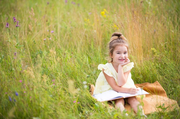 Une fille en robe jaune est assise dans l'herbe sur une couverture dans un champ et lit un livre en papier. Journée internationale des enfants. Heure d'été, enfance, éducation et divertissement, noyau de chalet. Espace de copie