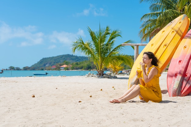 Fille en robe jaune sur une plage de sable tropicale travaille sur un ordinateur portable près de kayaks et boit de la mangue fraîche. Travail à distance, indépendant à succès. Fonctionne en vacances.