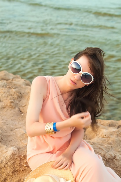 Fille en robe avec des lunettes et un chapeau assis sur une pierre au bord de la mer