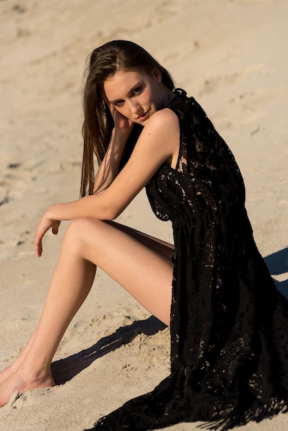 Une fille en robe noire est assise sur la plage dans le sable.