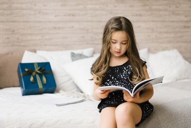 Une fille en robe noire tient un magazine et le lit, assise sur le lit, sur laquelle se trouve une boîte cadeau bleue avec un arc vert