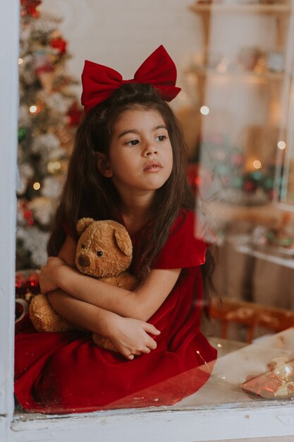 Fille en robe rouge avec un arc sur la tête avec un ours en peluche attend le Père Noël à la fenêtre