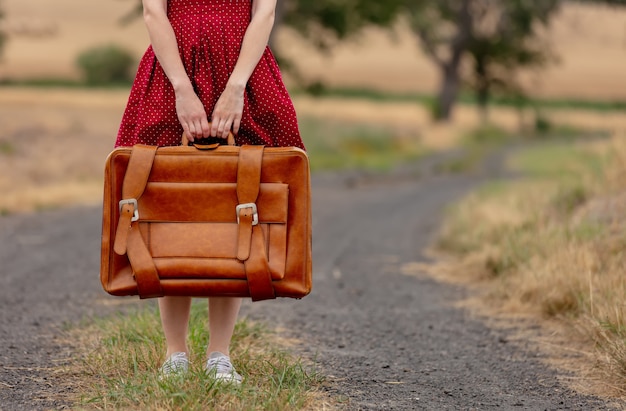 Fille en robe rouge avec une valise sur une route rurale avant la pluie