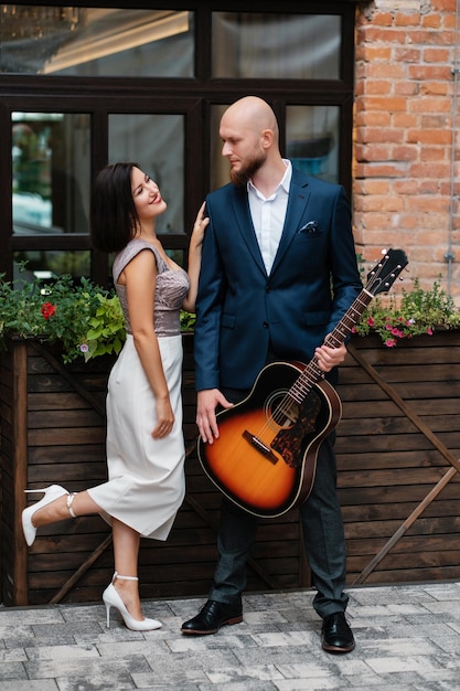 Une fille en robe de soirée embrasse un musicien Homme chauve barbu avec une guitare