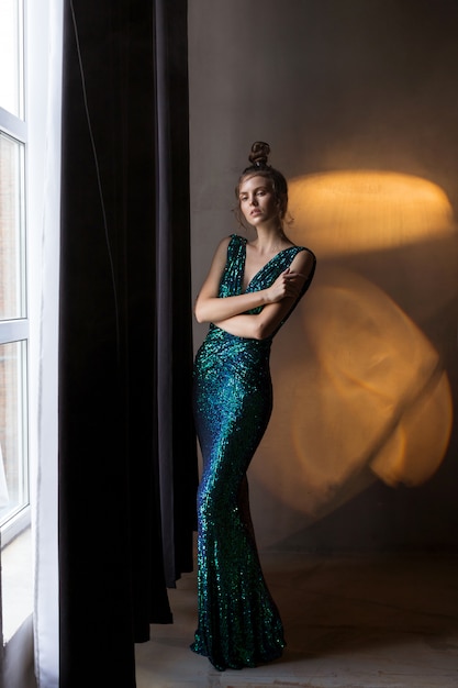 Fille en robe vert-bleu brillant se tient dans une lumière chaude, fumée, sombre