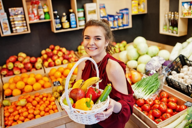 Fille en rouge tenant différents fruits et légumes au panier sur le magasin de fruits