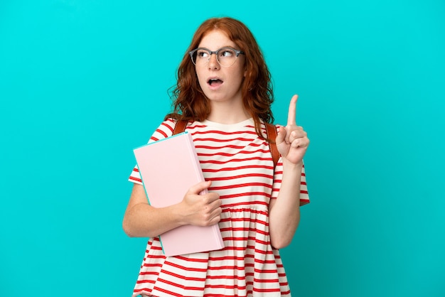 Fille rousse adolescente étudiante isolée sur fond bleu pensant à une idée pointant le doigt vers le haut