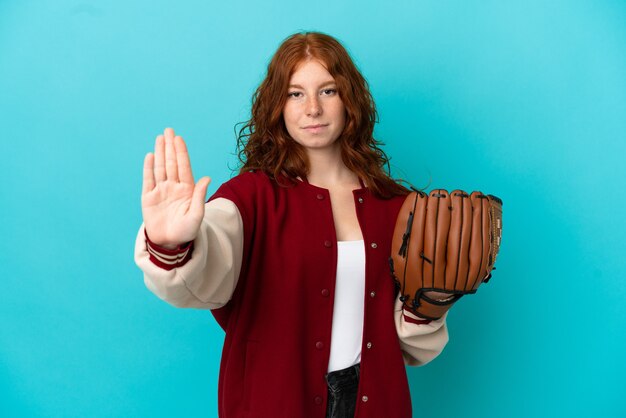 Fille rousse adolescente avec gant de baseball isolé sur fond bleu faisant un geste d'arrêt