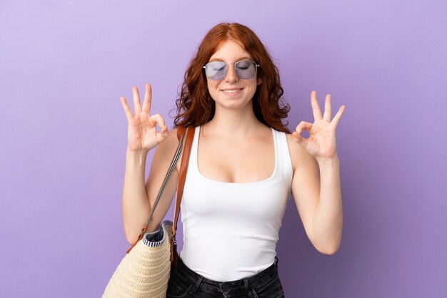 Fille rousse adolescente tenant un sac de plage isolé sur fond violet dans une pose zen