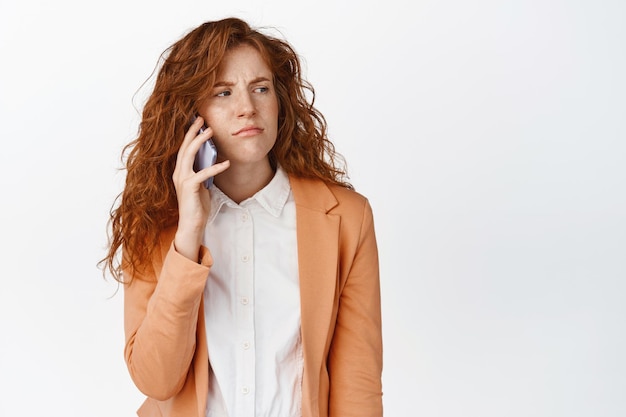 Fille rousse agacée appelant quelqu'un en attente d'une réponse sur smartphone fronçant les sourcils et grimaçant debout sur fond blanc