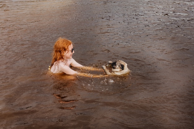 Une fille rousse apprend à son carlin à nager dans la rivière.