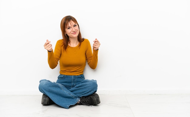 Fille rousse assise sur le sol isolé sur fond blanc faisant un geste d'argent