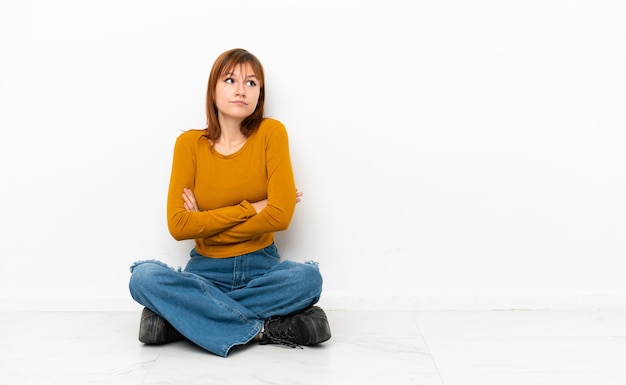 Fille rousse assise sur le sol isolée sur fond blanc faisant un geste de doute tout en soulevant les épaules