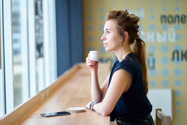 Fille rousse blanche en chemisier bleu foncé, boire du café au café et regarder à la fenêtre