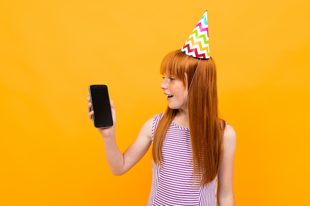 Fille rousse avec un bonnet de fête sur la tête tient le téléphone avec l'écran vers l'avant avec une mise en page sur fond jaune