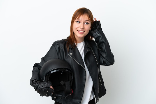 Fille rousse avec un casque de moto isolé sur fond blanc écoutant quelque chose en mettant la main sur l'oreille