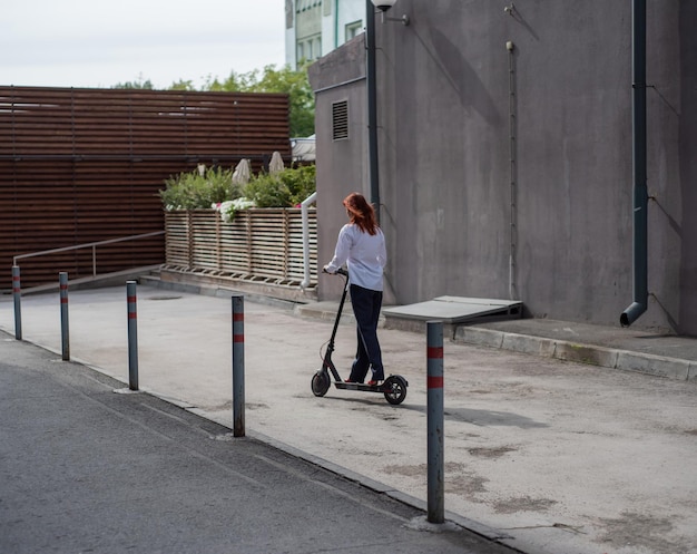 Une fille rousse en chemise blanche conduit un scooter électrique le long du mur Une femme d'affaires en tailleur-pantalon et talons hauts rouges se promène dans la ville dans une voiture moderne Femme d'affaires sur un scooter