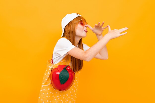 Fille rousse dans des verres et avec pomme rouge grimace à la caméra sur un fond jaune