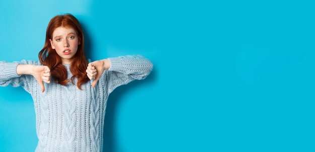 Fille rousse ennuyée et sceptique montrant les pouces vers le bas à l'air indifférent et indifférent debout sur