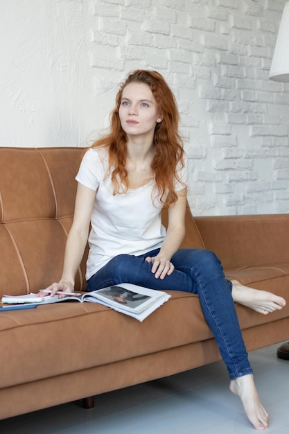 Une fille rousse est allongée sur le canapé et lit un magazine à la maison