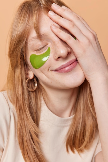 Une fille rousse heureuse avec une peau rougie applique des patchs de collagène gree sous les yeux fait que la paume du visage garde les yeux fermés sourit doucement subit des procédures de beauté pose à l'intérieur Concept de soins de la peau