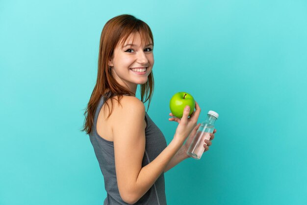 Fille rousse isolée sur fond bleu avec une pomme et avec une bouteille d'eau