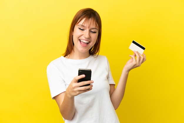 Fille rousse isolée sur fond jaune achetant avec le mobile avec une carte de crédit