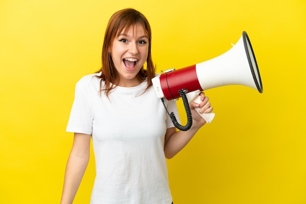 Fille rousse isolée sur fond jaune tenant un mégaphone et avec une expression surprise