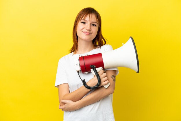 Fille rousse isolée sur fond jaune tenant un mégaphone et souriant