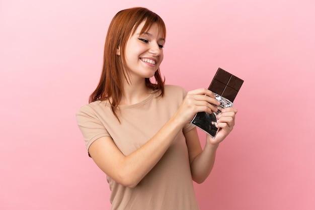 Fille rousse isolée sur fond rose prenant une tablette de chocolat et heureuse