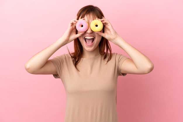 Fille rousse isolée sur fond rose tenant des beignets dans un œil