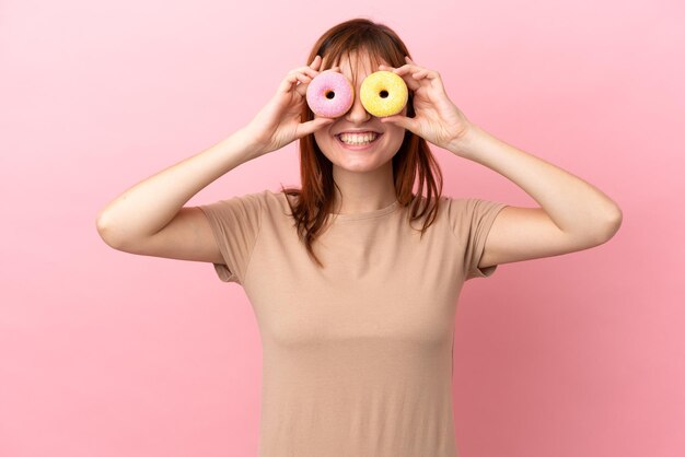 Fille rousse isolée sur fond rose tenant des beignets dans les yeux