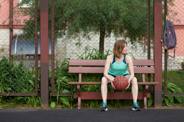 Fille rousse maigre de race blanche avec ballon dans les mains, assise et attendant un match sur le terrain de basket