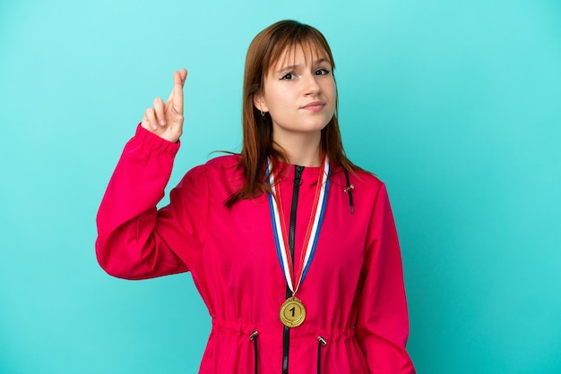 Fille rousse avec des médailles isolées sur fond bleu avec les doigts croisés et souhaitant le meilleur