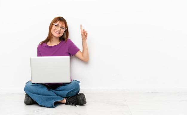 Fille rousse avec un ordinateur portable assis sur le sol montrant et levant un doigt en signe du meilleur