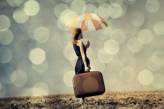 Fille rousse avec parapluie et valise au champ venteux