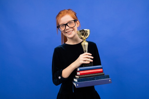 Fille rousse avec une pile de livres et une coupe de la victoire sur un fond bleu studio.