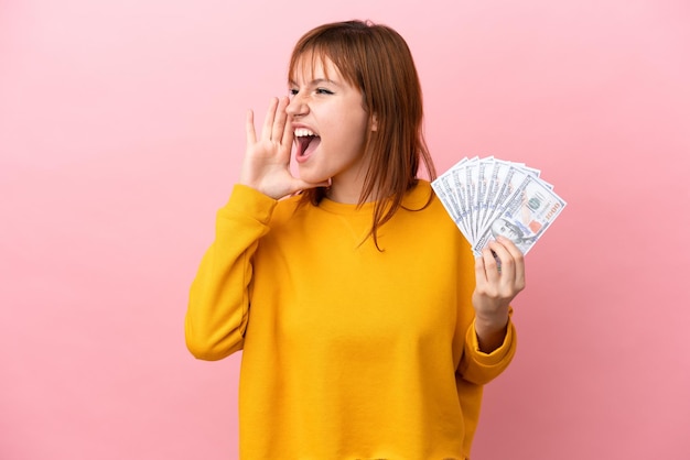 Fille rousse prenant beaucoup d'argent isolé sur fond rose criant avec la bouche grande ouverte sur le côté