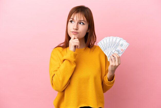 Fille rousse prenant beaucoup d'argent isolé sur fond rose et levant les yeux