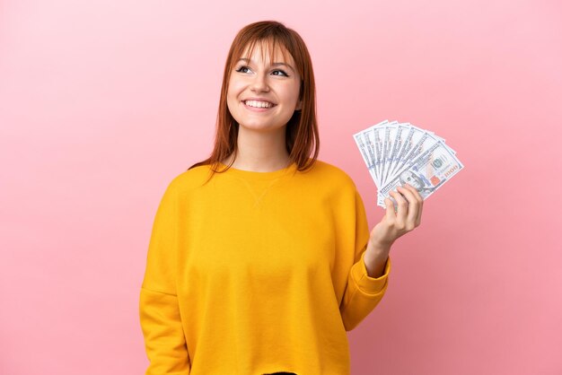 Fille rousse prenant beaucoup d'argent isolée sur fond rose en pensant à une idée tout en levant les yeux