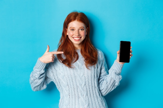 Fille rousse satisfaite pointant sur l'écran du téléphone, montrant une application pour smartphone ou une promotion en ligne et souriante, debout en pull sur fond bleu.