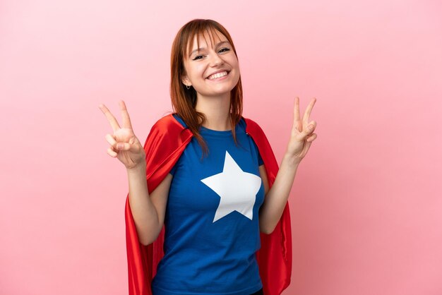 Fille rousse super héros isolée sur fond rose montrant le signe de la victoire avec les deux mains