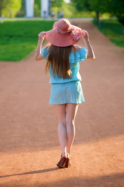 Fille rousse, vêtue d'une robe bleue et d'un chapeau de couleur corail, se promène dans le parc d'été, vue de l'arrière.